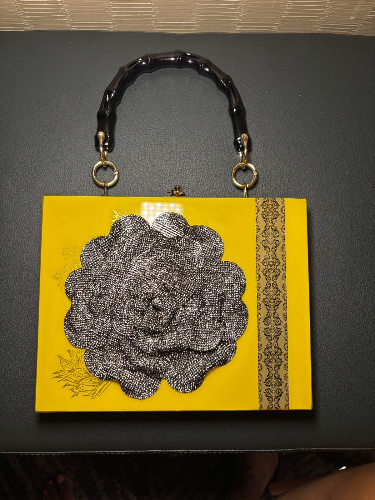 A bright yellow clutch handbag with a black faux-bamboo handle, gold-tone hardware, and a large black floral applique on the front, photographed on a dark surface.