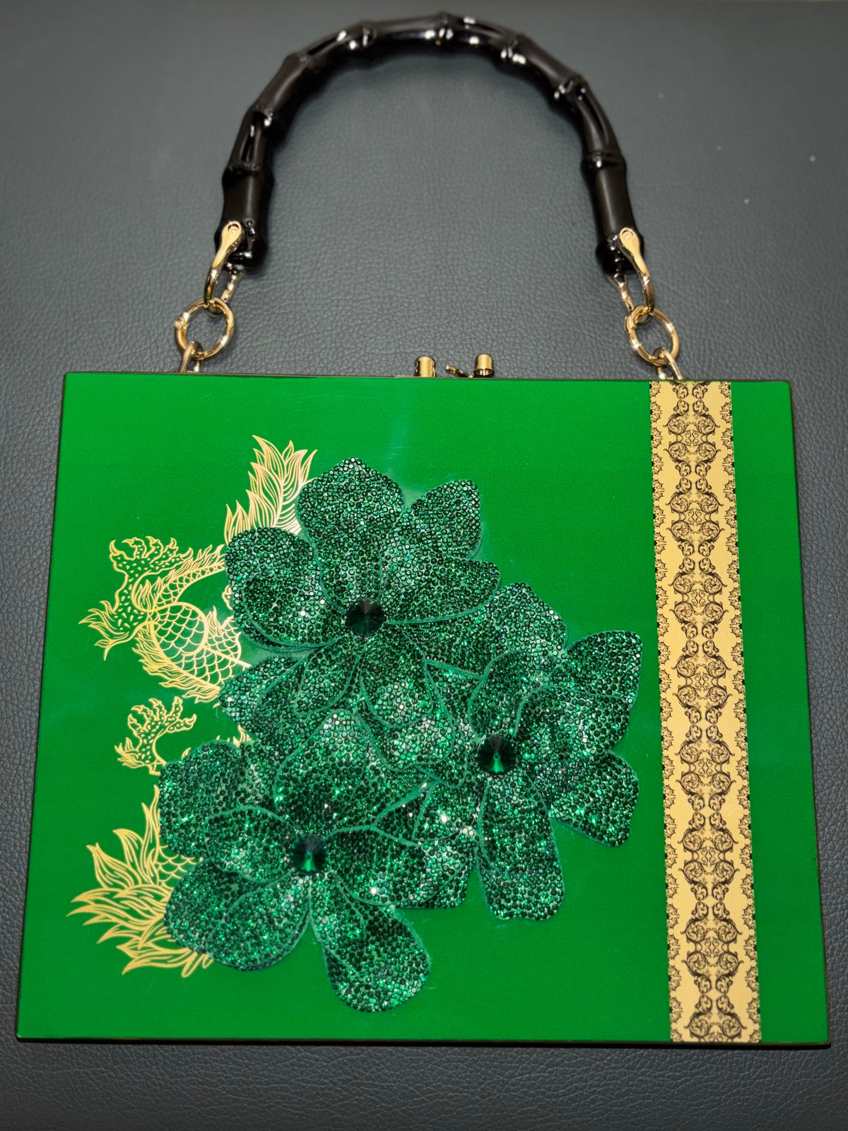 A green rectangular clutch handbag with a bamboo-style black handle, gold-tone hardware, a large green floral applique on the front, and a vertical gold patterned trim strip.