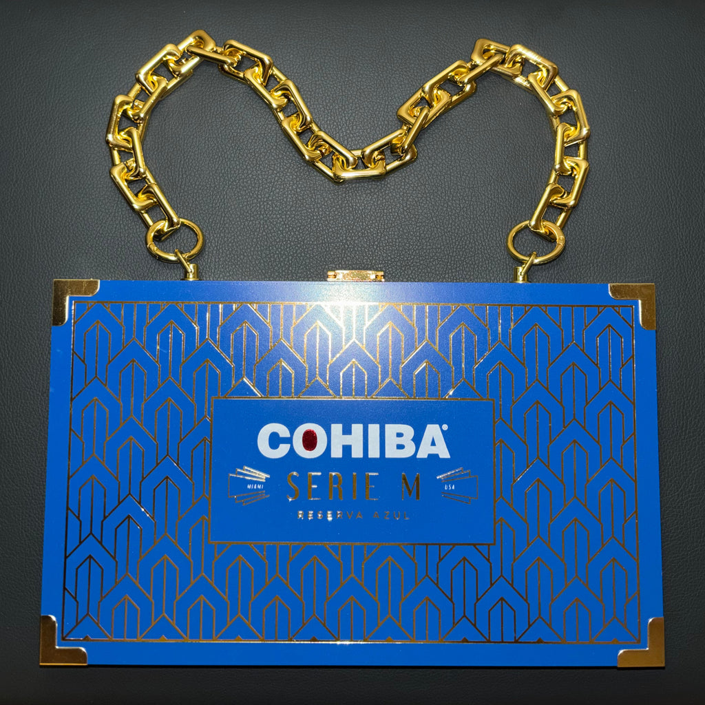A blue rectangular clutch bag with gold geometric patterns, a gold chain shoulder strap, and gold corner accents. The front features a blue label with the text 'COHIBA SERIE M'.