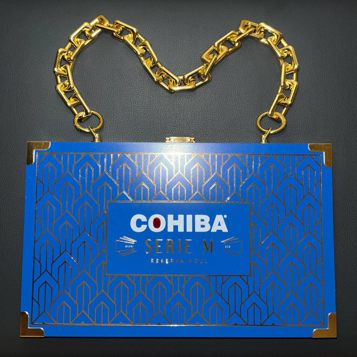 A blue rectangular clutch bag with gold geometric patterns, a gold chain shoulder strap, and gold corner accents. The front features a blue label with the text 'COHIBA SERIE M'.