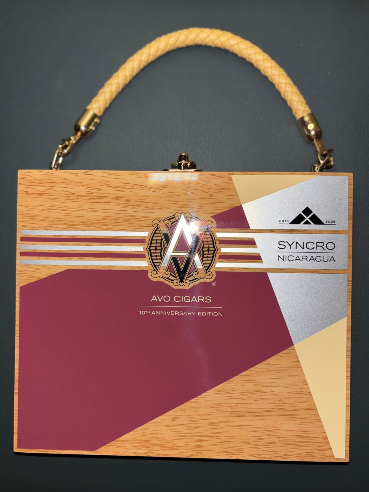 A rectangular AVO Cigars 'Syncro' 10th Anniversary Edition handbag with a wood-grain patterned front, maroon and beige geometric panels, a gold clasp, and a braided gold handle, set against a dark gray background.