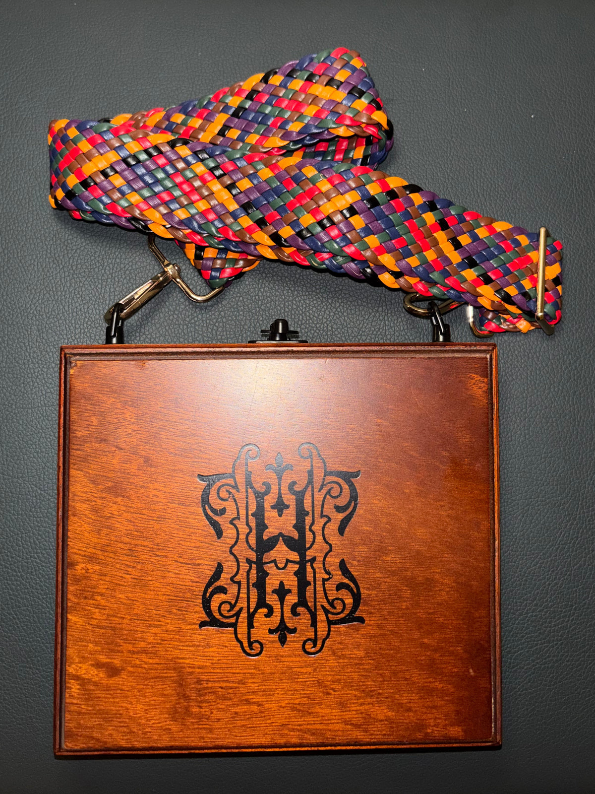 A square wooden case with a warm brown wood-grain finish and an engraved black monogram, featuring a colorful woven strap with red, blue, yellow, and purple threads, displayed on a dark gray textured surface.